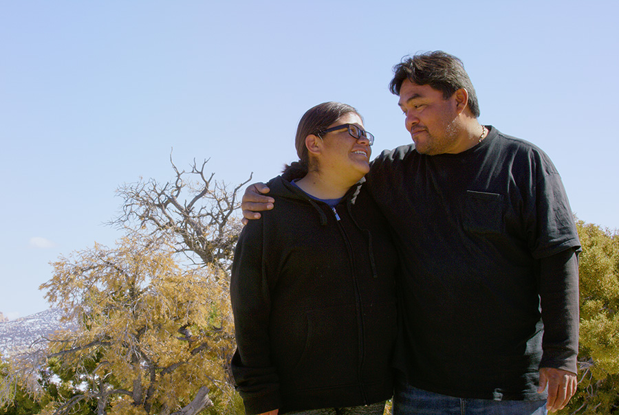 Native American couple with their arms around each other smile at each other on a sunny day outdoors.